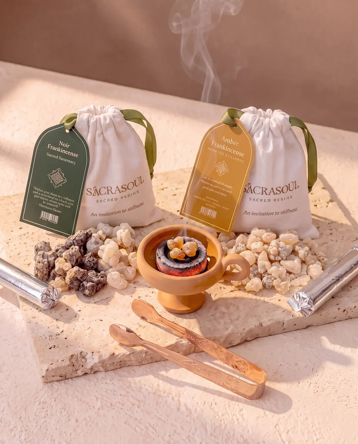 De First Flame-kit op travertijn: Sacrasoul-zakjes met labels Noir Frankincense en Amber Frankincense, ervoor de harsen opgehoopt, met de aangestoken terracottabrander, de roségouden tang en twee rollen kooltjes.