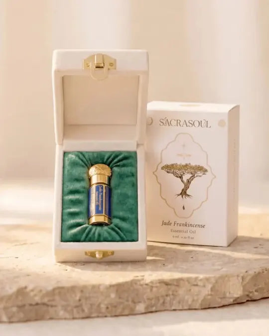 Het goud-blauwe flesje Jade-wierookolie in het open crèmekleurige fluwelen etui, naast het bedrukte doosje met de tekst Sacrasoul, Jade Frankincense Essential Oil, 6 ml.